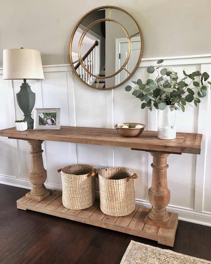 modern farmhouse entryway table with two legs decorated with baskets, flower arrangement and round mirror above on a panelled wall