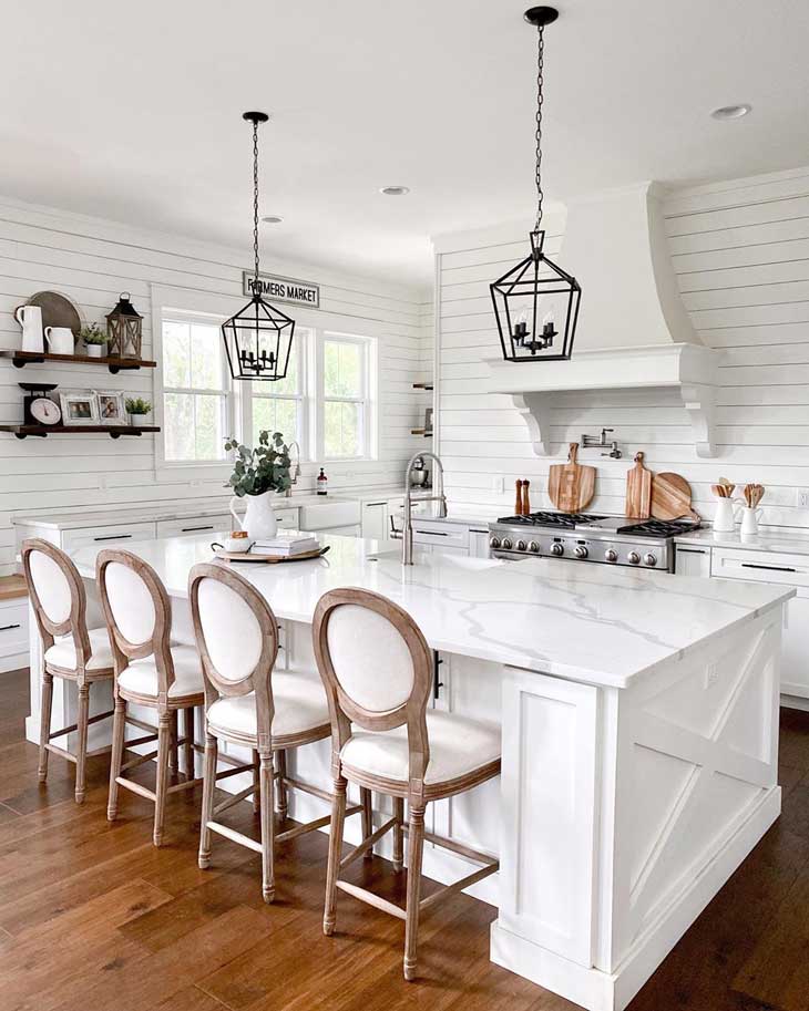 black pendant lights over white kitchen island with marble countertop and wood upholstered barstools in a kitchen with shiplap backsplash